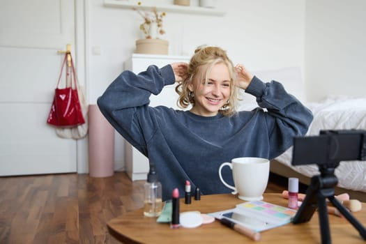 Portrait of cute blond girl, lifestyle blogger sits on floor with video camera and stabiliser, shows how to do hairstyle and makeup, does casual vlogging for social media, sits in room.