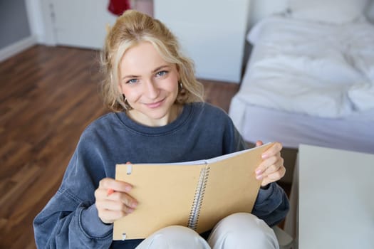 Portrait of smiling, charismatic young woman, writing down notes, making plans and putting it in planner, holding journal, sitting in bedroom and looking happy at camera.