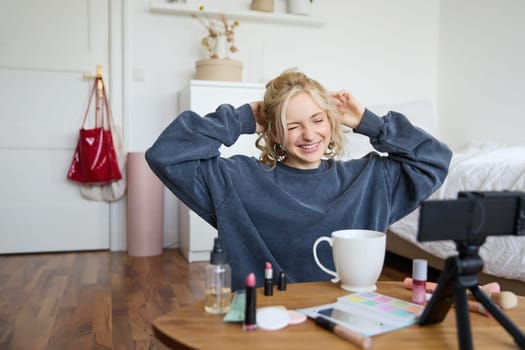 Portrait of cute smiling woman, blond girl drinks tea and records a casual, lifestyle video blog, vlogger sits in a room with camera and stabiliser, holds cup of coffee and talks.