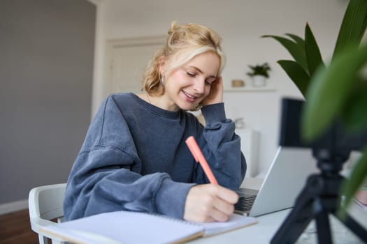 Cute smiling girl sits in a room, writes down notes, doing homework, records video of herself on digital camera, creates content for vlog, lifestyle blogger doing daily routine episode.