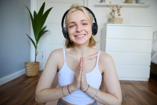 Portrait of woman meditating at home, sitting in headphones, listening to yoga podcast, holding hands in namaste sign.