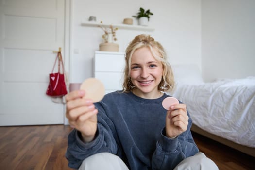 Image of smiling blond woman, vlogger, showing makeup products, beauty items, recommending to followers, sitting on floor in bedroom.