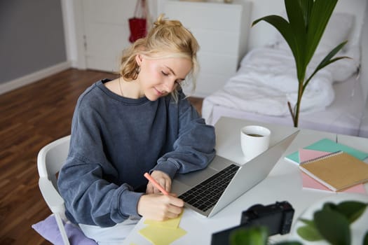 Portrait of smiling young woman, female student, doing distance learning course, using laptop, studying at home, writing down, making notes.