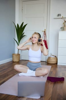 Vertical shot of smiling young woman using video tutorials for workout at home, sitting with laptop in wireless headphones on yoga mat, follows fitness instructor on social media, repeats exercises.