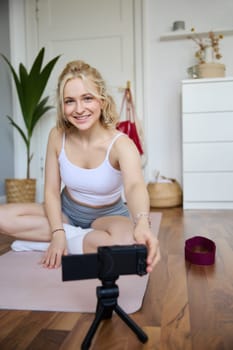 Portrait of young woman personal yoga instructor, recording workout video at home, using digital camera to vlog her exercises, using rubber mat.