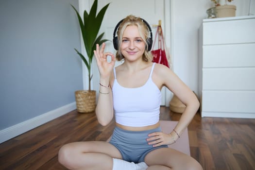 Portrait of young smiling beautiful woman, wearing wireless headphones, working out at home, sitting on rubber yoga mat, showing okay sign, satisfied with exercises.