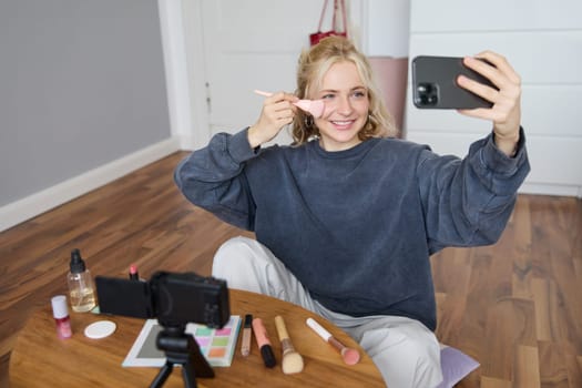 Image of stylish young woman, social media influencer, taking pictures on mobile phone, doing makeup tutorial for followers online, recording video vlog in her bedroom, showing brush.