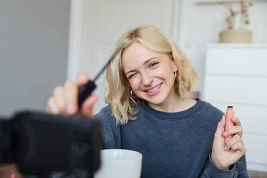 Smiling young beautiful lifestyle blogger, girl records a beauty vlog in her room, using professional video camera, showing makeup cosmetics to her followers, creates content for social media.