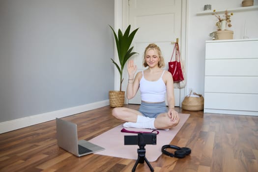 Portrait of young female athlete, fitness trainer recording vlog, training session on digital camera, sitting in a room on rubber yoga mat, showing exercises.