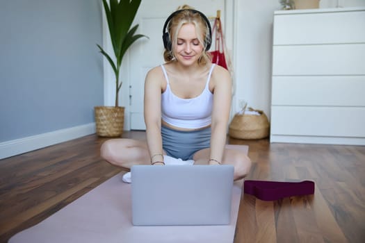 Portrait of young sporty woman following online video instructions during fitness workout, using laptop, sitting on yoga mat.