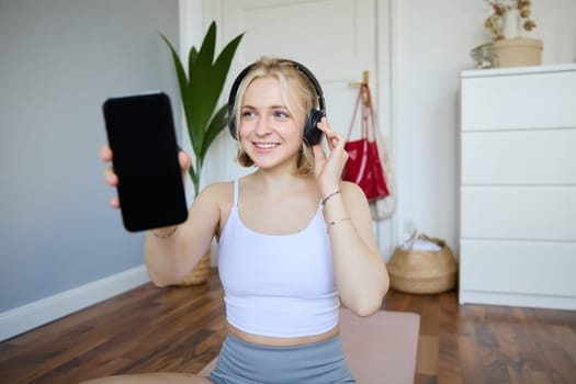 Portrait of fit and healthy woman workout at home, wearing headphones, showing smartphone screen, empty dark display. Wellbeing and lifestyle concept