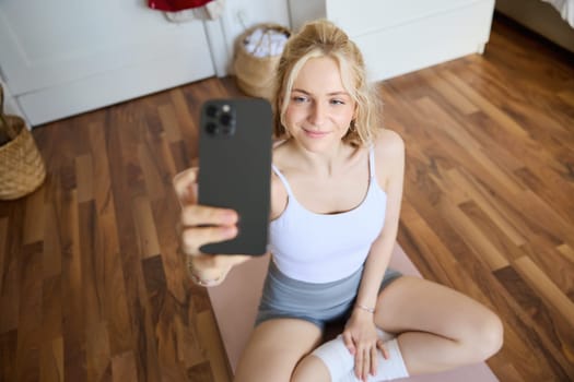 Portrait of beautiful young fitness trainer, showing home workout, taking selfie on mobile phone while sitting on yoga rubber mat in bedroom, wearing activewear.