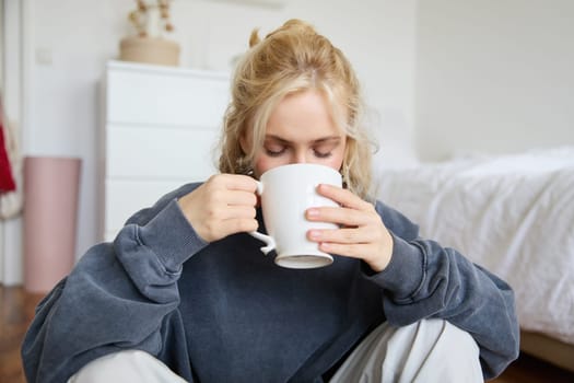 Portrait of young woman sitting on bedroom floor, drinking tea, holding white mug and smiling at camera.