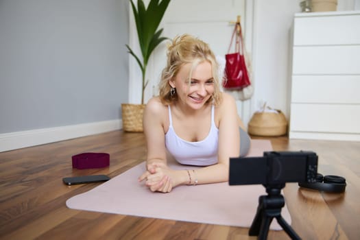 Portrait of young fitness instructor, vlogger creating content at home, doing workout and record exercises on digital camera, using rubber yoga mat.