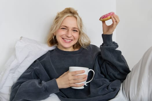 Close up portrait of beautiful smiling, blond woman, lying in bed, drinks tea and eats doughnut on a lazy weekend or day-off.