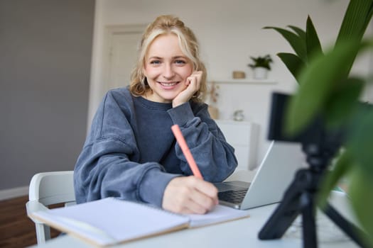 Cute smiling girl sits in a room, writes down notes, doing homework, records video of herself on digital camera, creates content for vlog, lifestyle blogger doing daily routine episode.