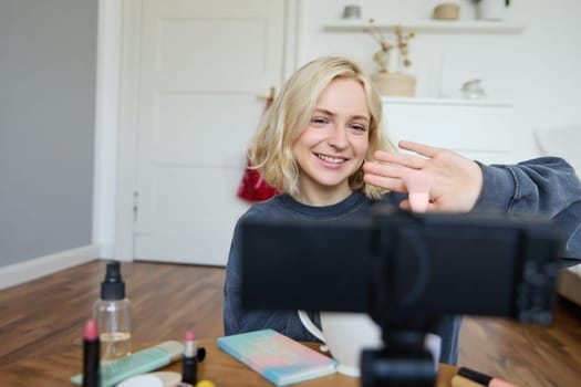 Close up portrait of happy young beauty blogger, records lifestyle vlog in her room, using camera with stabiliser, shows makeup brush and cosmetics.