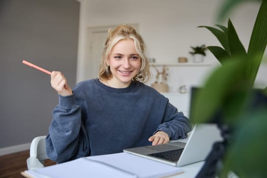 Portrait of young charismatic woman, smiling, holding pen, looking at laptop, video chats, doing online course, answering a question, studying with tutor.