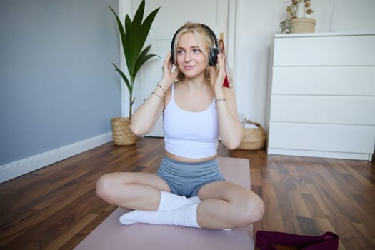 Portrait of happy, beautiful athletic woman, working out at home, listening music to boost energy while active training session, using wireless headphones and rubber yoga mat.