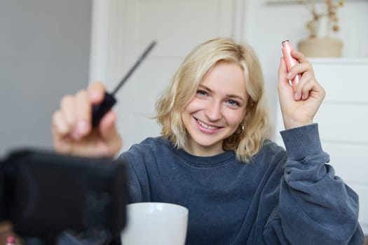 Portrait of young smiling woman in her room, recording video on camera, lifestyle vlog for social media, holding mascara, reviewing her makeup beauty products, showing how to use cosmetics.