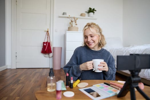 Portrait of cute blond woman, video content maker, vlogger recording on digital camera in bedroom, drinking cup of tea and talking to audience.