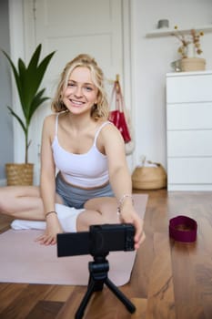 Vertical shot of athletic, fitness woman recording video of herself on digital camera, creating content for social media about workout and home exercises.