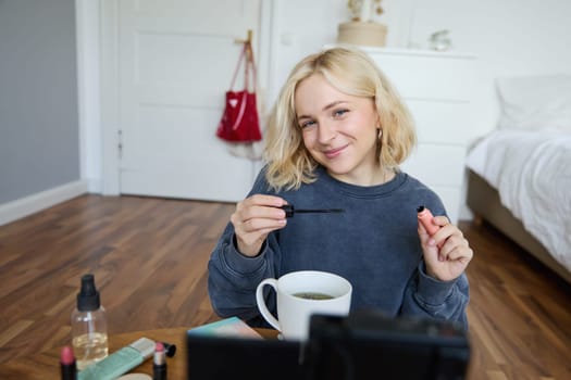 Portrait of cute lifestyle blogger sits in her room in front of video camera, recording for social media account, putting on makeup, reviewing beauty products, holding mascara.