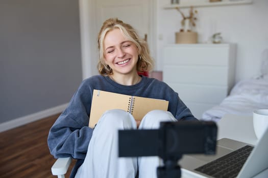 Portrait of blond smiling woman, records video on digital camera how she writes in notebook, talks to followers, doing lifestyle blog content in her room.