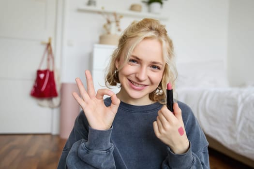 Portrait of young blond woman, content creator, recording video for social media about makeup, showing okay hand sign and lipstick, recommending good cosmetic product.