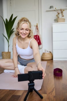 Portrait of young fitness instructor, vlogger creating content at home, doing workout and record exercises on digital camera, using rubber yoga mat.