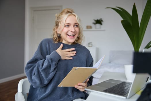 Portrait of smiling, beautiful young blond woman, student working on assignment from home, online learning in her bedroom, talking to video camera, chatting, holding notebook.