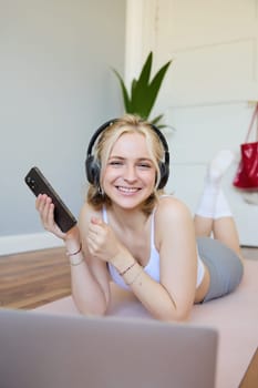 Vertical shot of young blond woman in headphones, lying on rubber mat, using laptop, holding mobile phone, listens to music or workout podcast, doing exercises at home.