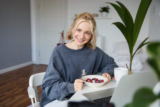 Portrait of smiling blond young woman, eating in front of laptop, watching videos online while having breakfast, enjoying dessert, sitting in bedroom.