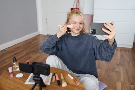 Portrait of beautiful blond girl doing makeup on camera, recording vlog using digital camera and live stream on mobile phone app, chatting with audience.