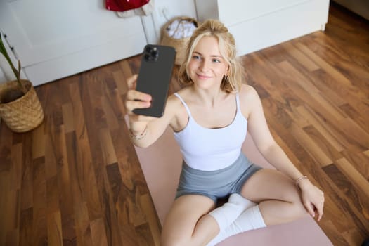 Portrait of stylish, young beautiful woman takes selfie while working out at home, using smartphone to make photos on yoga mat, posing in fitness clothes.