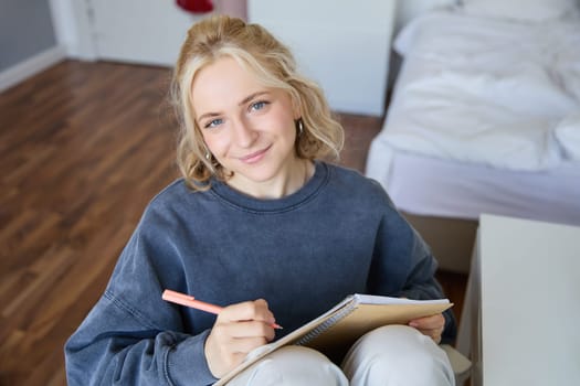 Close up portrait of beautiful young woman, writing in her diary, journal or planner, sitting in a room, smiling and looking at camera.