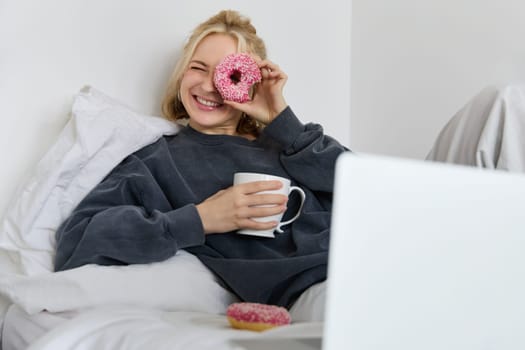 Portrait of happy cute woman, lying in bed with laptop, drinking tea, looking through doughnut hole and smiling at camera.