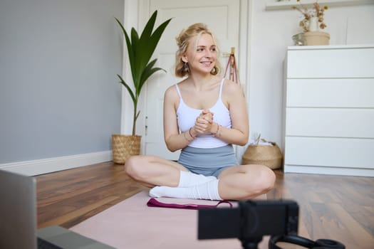 Portrait of young fitness instructor, vlogger showing exercises on camera, recording herself, sitting on mat with laptop, doing workout, explaining yoga movements to followers.