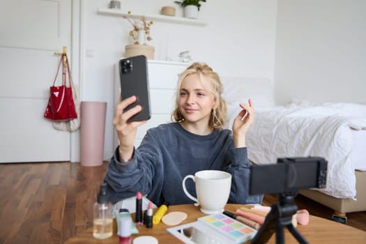 Portrait of young beauty blogger, recording video on digital camera, doing online live stream while applying makeup, talking to her followers, sitting on bedroom floor.