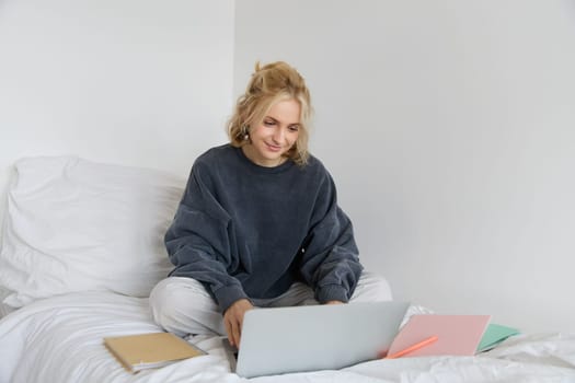 Image of happy young woman, student e-learning from home, connect to online course on her laptop, sits on bed with notebooks, smiling and looking happy.