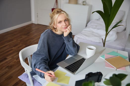 Portrait of young thoughtful girl, studying, making notes, writing down something in notebook, doing homework in her room, sitting in front of laptop, frowning while thinking.