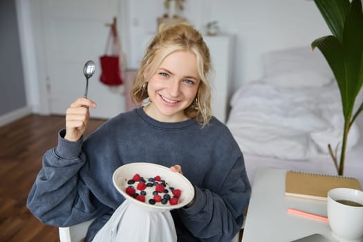 Portrait of smiling blond woman, eating breakfast, holding bowl and spoon, sitting in bedroom, looking happy at camera. Lifestyle concept