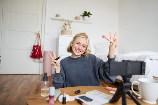 Portrait of young blond woman, teenage girl records video for her social media account, shows makeup on camera, recommends lipstick to online followers, creates content in her room.