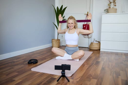 Portrait of young athletic woman recording home workout video, shooting content for sport fitness vlog, using resistance band and digital camera.