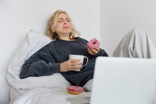 Portrait of sad and depressed woman, feeling heartbroken, lying in bed with comfort food, eating doughnut, crying from upsetting movie, watching something on laptop.