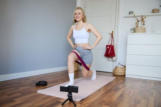 Portrait of fitness instructor, young sporty vlogger woman doing exercises on camera, shooting video about workout, using resistance band and doing sit-ups at home in empty room.