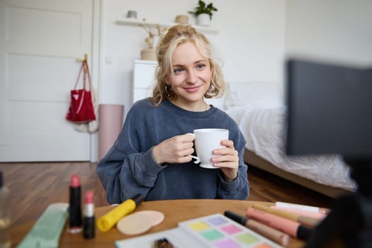 Portrait of cute blond woman, video content maker, vlogger recording on digital camera in bedroom, drinking cup of tea and talking to audience.