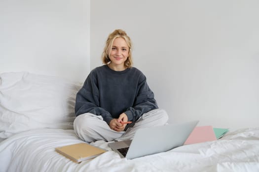 Portrait of happy blond woman, freelancer working from home, sitting on bed with laptop and notebooks. Student doing homework in bedroom, connects to online class via video chat.