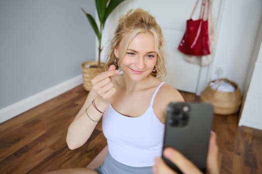 Portrait of smiling, beautiful young woman, social media content creator, takes selfies on smartphone during workout, doing exercises at home on yoga mat.