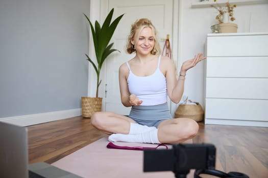 Portrait of beautiful fitness blogger, recording video on digital camera, showing workout exercises, explaining fitness movements or yoga training to followers, sits on rubber mat.